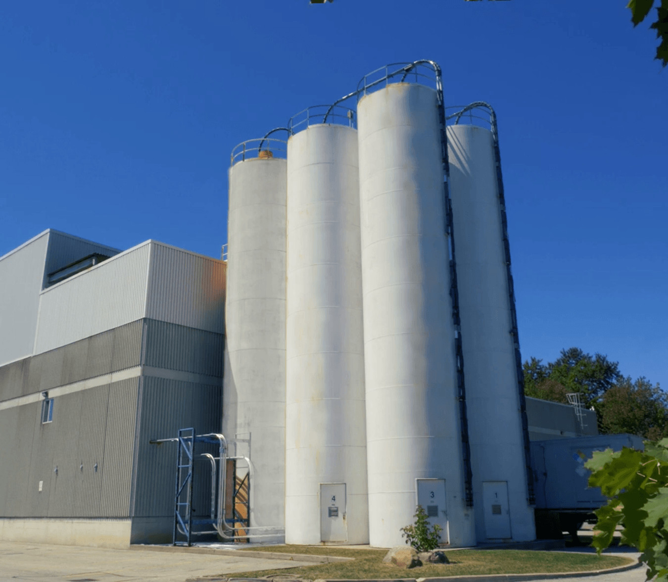 Four tall, white industrial silos are positioned next to a gray building under a clear blue sky; each silo is labeled with a number near its base.