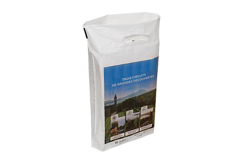 A white plastic bag with a handle features a blue label showing scenic landscapes and French text, "Trois Circuits de Grandes Découvertes.