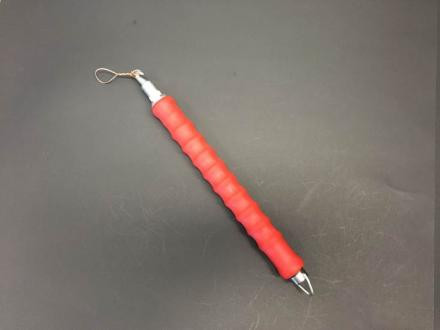 Sand/Soil Bags – a metal tool with a red grooved handle, a small wire loop at one end, and a pointed tip at the other, placed on a dark surface.