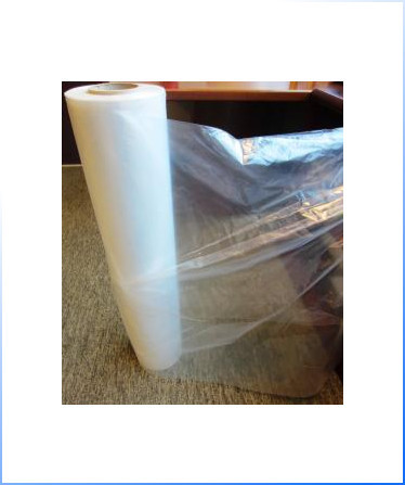 A large roll of clear plastic film is partially unrolled on a carpeted floor next to a dark-colored piece of furniture.