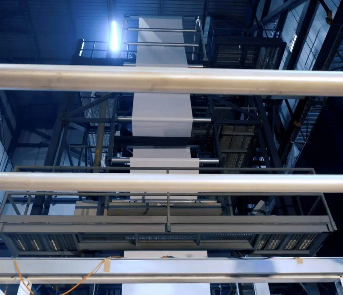 Large industrial printing press with stacked rolls of white paper and metal railings inside a factory setting.
